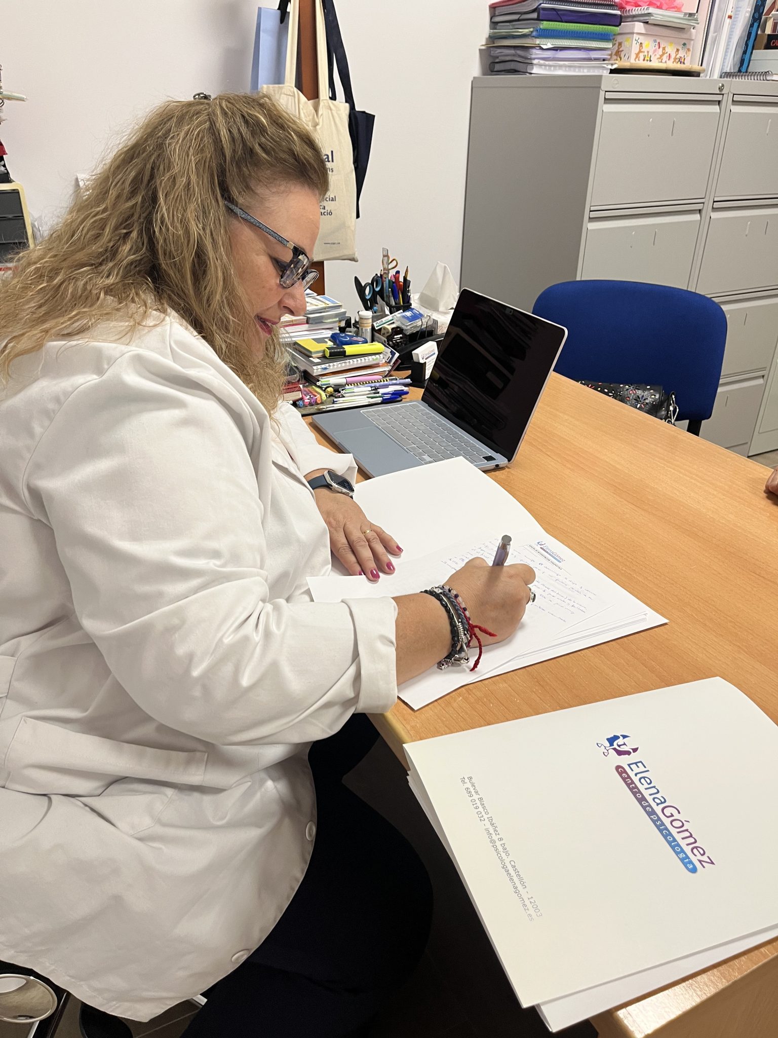 Mujer con bata firmando documentos en una oficina.
